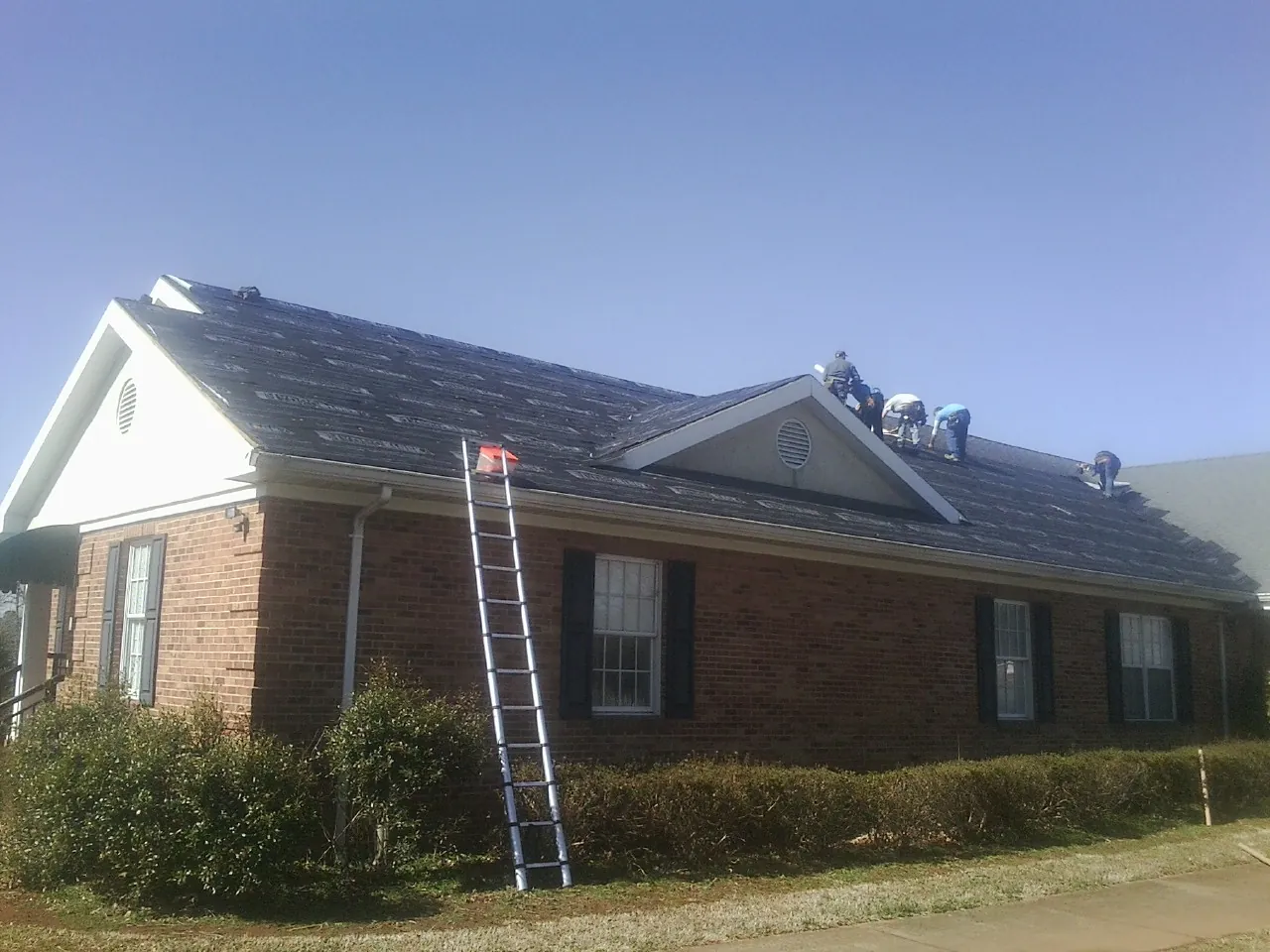 Crew tearing shingles off a brick ranch roof with a ladder set — Emergency Roof Repair work by our crew in Evans Mills