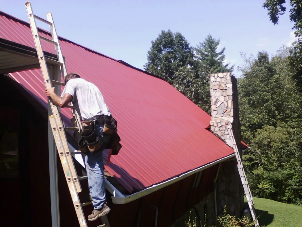 Roofer on a ladder beside a red metal roof and stone chimney — Soffit & Fascia Repair by Roofing Brothers in Evans Mills